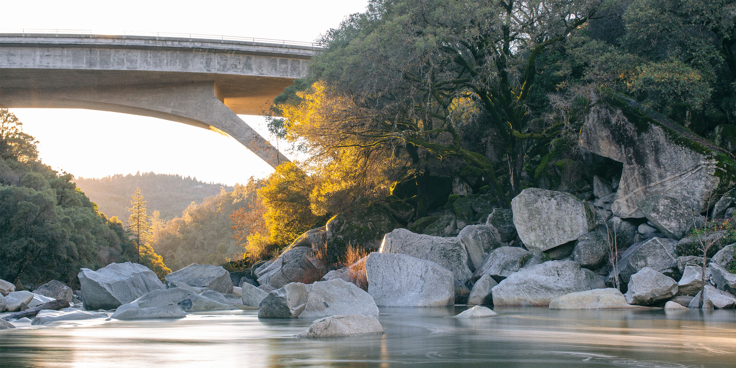 Long-exposure photo of the South Yuba River at golden hour, showing the water flowing around rocks and under a bridge, with trees on the right side bank