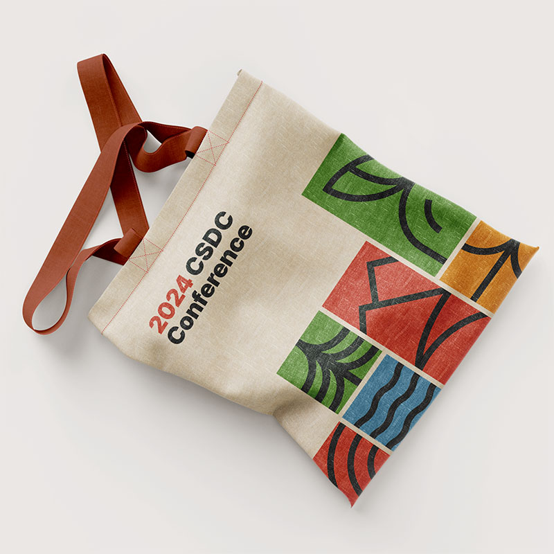 A canvas tote bag with the text 2024 CSDC Conference and the colorful graphics from the brand identity printed on it. It is set against a gray background.