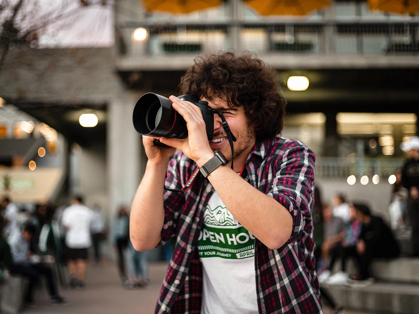 A photo of me smiling behind my camera against a blurry bokeh background. I'm wearing a red flannel and a white Open House 2020 shirt.